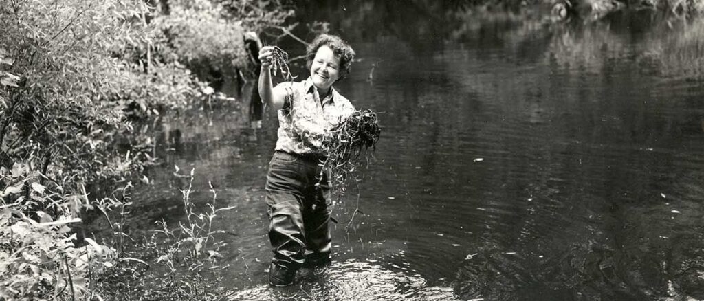 Historic photo of scientist Ruth Patrick smiling while taking samples and standing in a waterway.