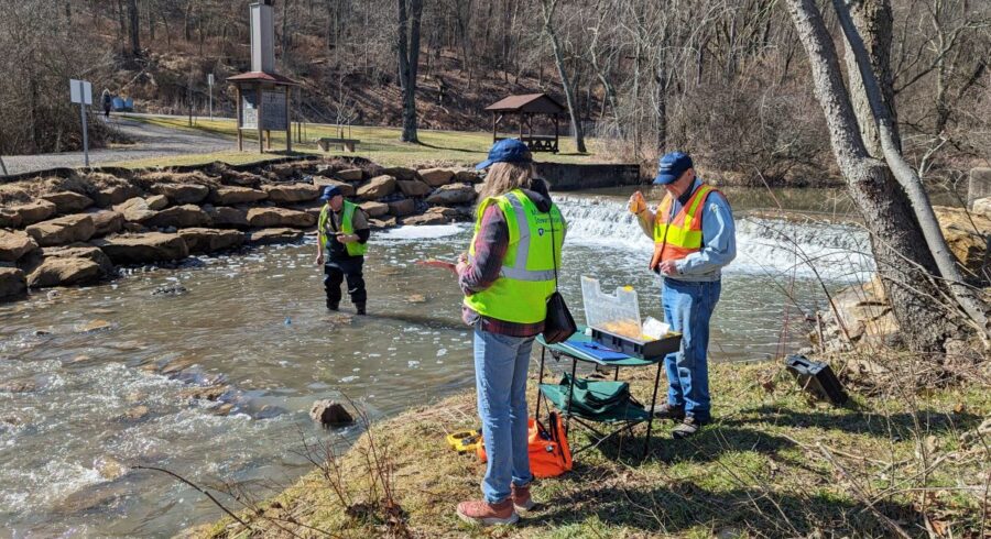 Three people stand in and around creek taking water samples to study.