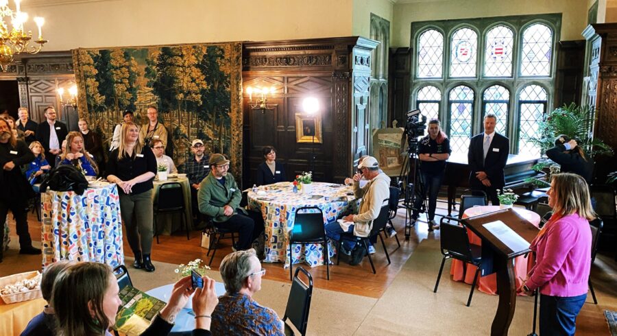 Woman speaking at podium inside with a group of people around listening.