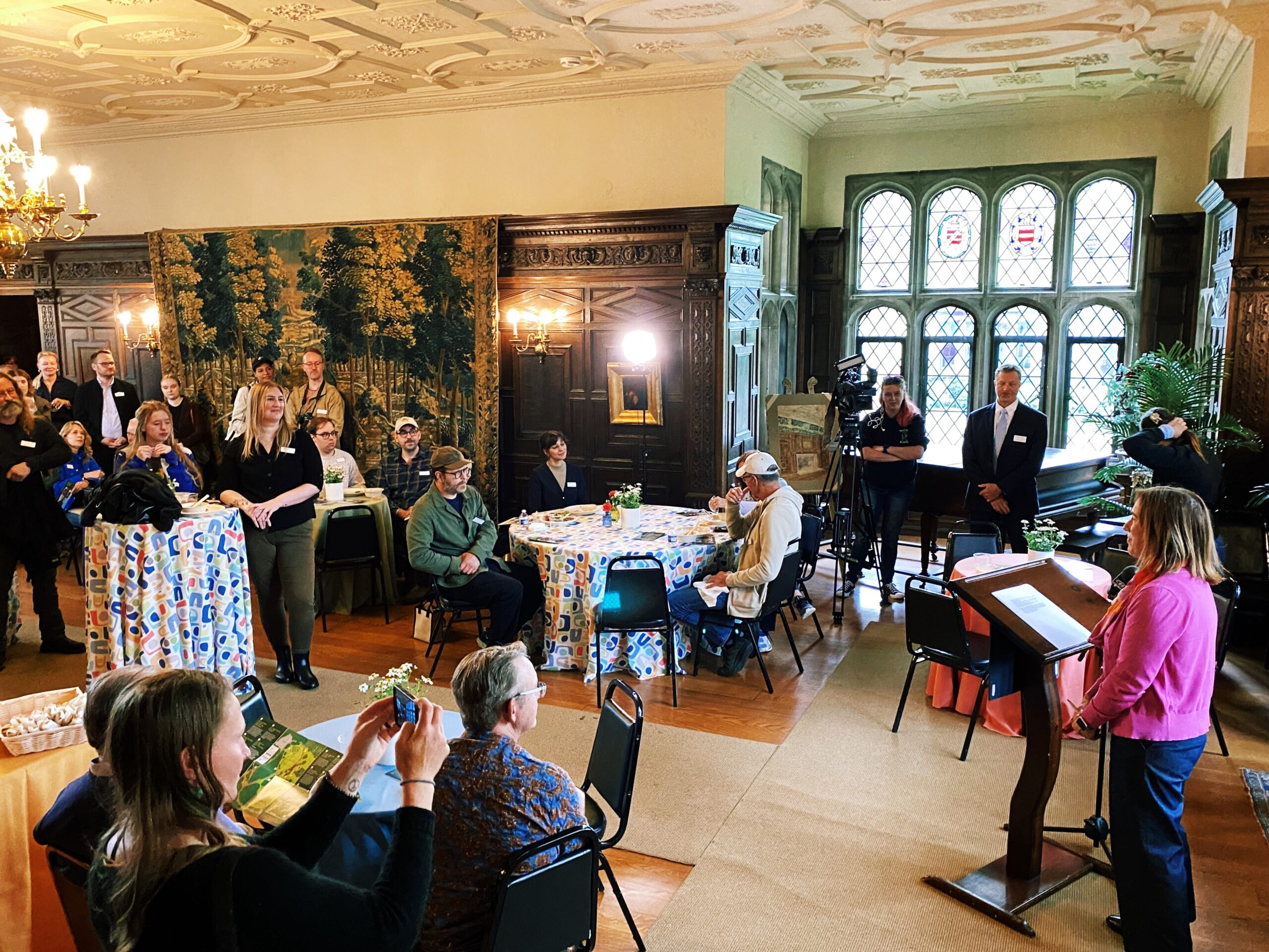Woman speaking at podium inside with a group of people around listening.
