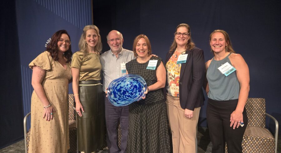 Group of people standing and smiling on stage. Person in middle is holding blue bowl award.
