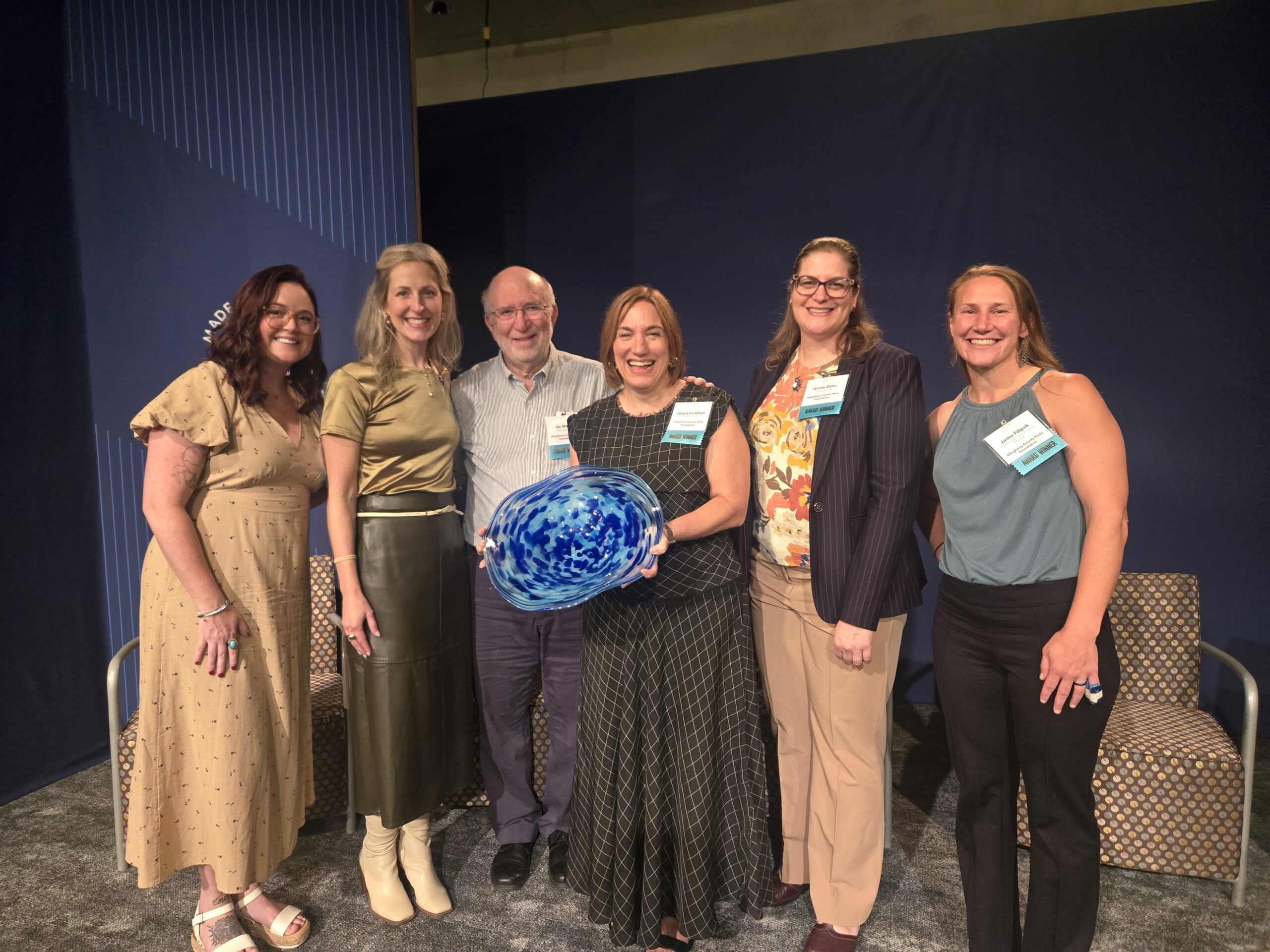 Group of people standing and smiling on stage. Person in middle is holding blue bowl award.